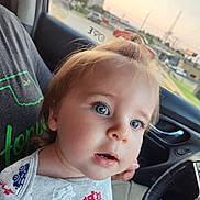 Kenneth is registered to the contest to win money with this photo: toddler, child, blue_eyes, topknot, shirt, fire_truck_pattern, car_interior, steering_wheel, person, gray_shirt, green_text, window, outside_view, sunset, vehicle, seat, hand, curious_expression, close_up, portrait