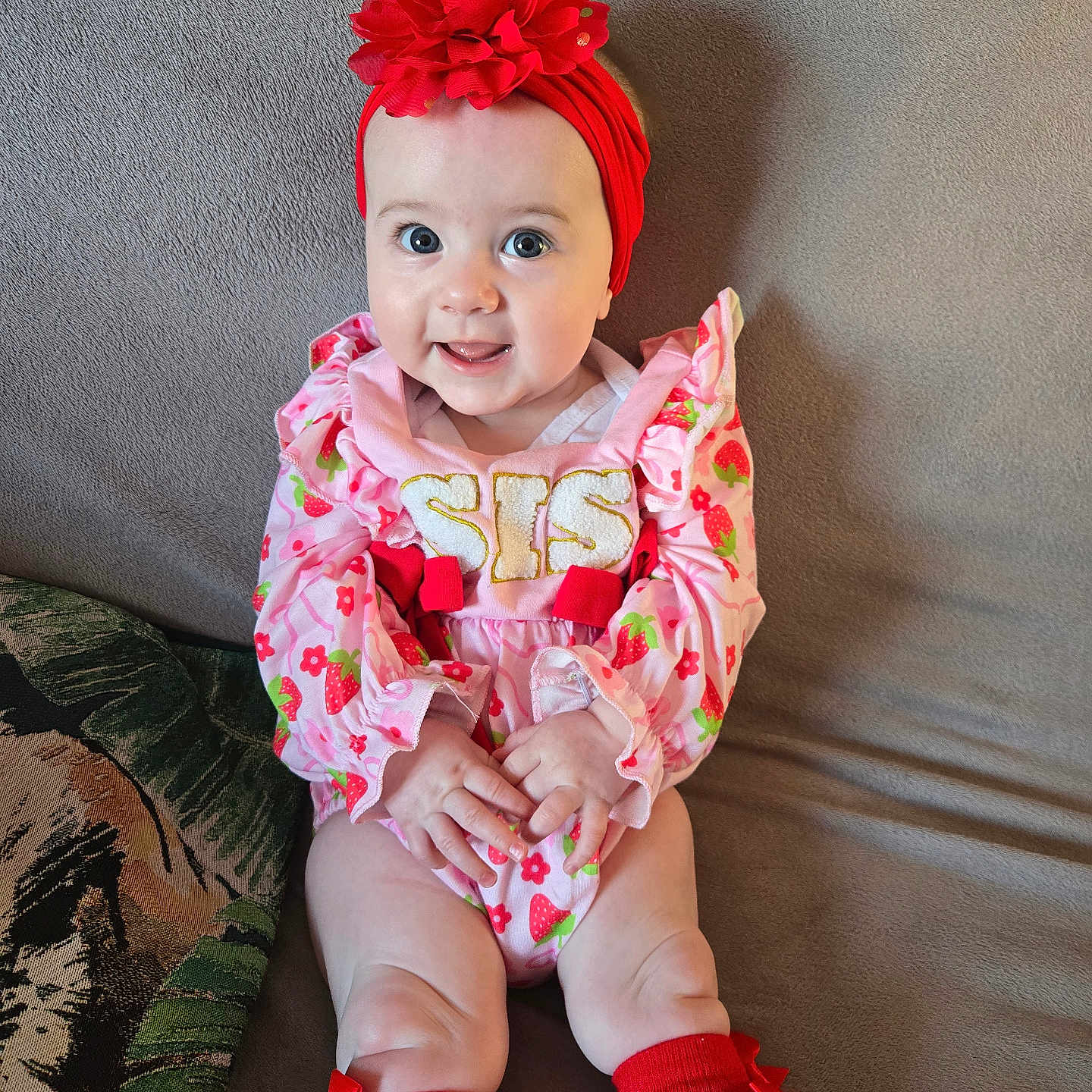 Leila-Mae is registered to the contest to win money with this photo: baby, child, clothing, couch, cute, face, flower, hands, happy, headwear, indoor, infant, pillow, pink_outfit, portrait, red_headband, red_socks, sitting, smiling, strawberry_pattern