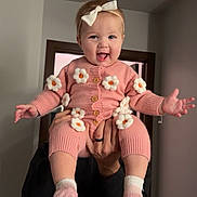 Lenora is registered to the contest to win money with this photo: baby, child, pink_clothing, knitted_outfit, flowers, headband, bow, socks, indoor, adult_hands, happy, playful, tongue_out, person, holding, smile, cute, infant, portrait, warm_clothing