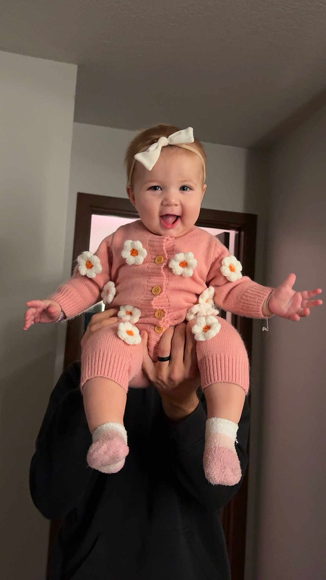 Lenora is registered to the contest to win money with this photo: baby, child, pink_clothing, knitted_outfit, flowers, headband, bow, socks, indoor, adult_hands, happy, playful, tongue_out, person, holding, smile, cute, infant, portrait, warm_clothing