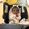 baby, infant, car_seat, headband, bottle, blanket, clothing, indoors, person, child, cute, curly_hair, sitting, feeding, toy, pink, yellow, black, white, floor