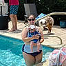 woman, baby, swimming_pool, sunglasses, dog, man, towel, swimsuit, sunhat, poolside, outdoor, summer, family, water, greenery, baby_seat, paved_floor, pet, sunny, relaxation