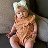 baby, infant, child, headband, bow, romper, orange_clothing, couch, cushion, soft_texture, seated, indoors, portrait, cute, small_hands, legs, face, expression, looking_at_camera, comfortable