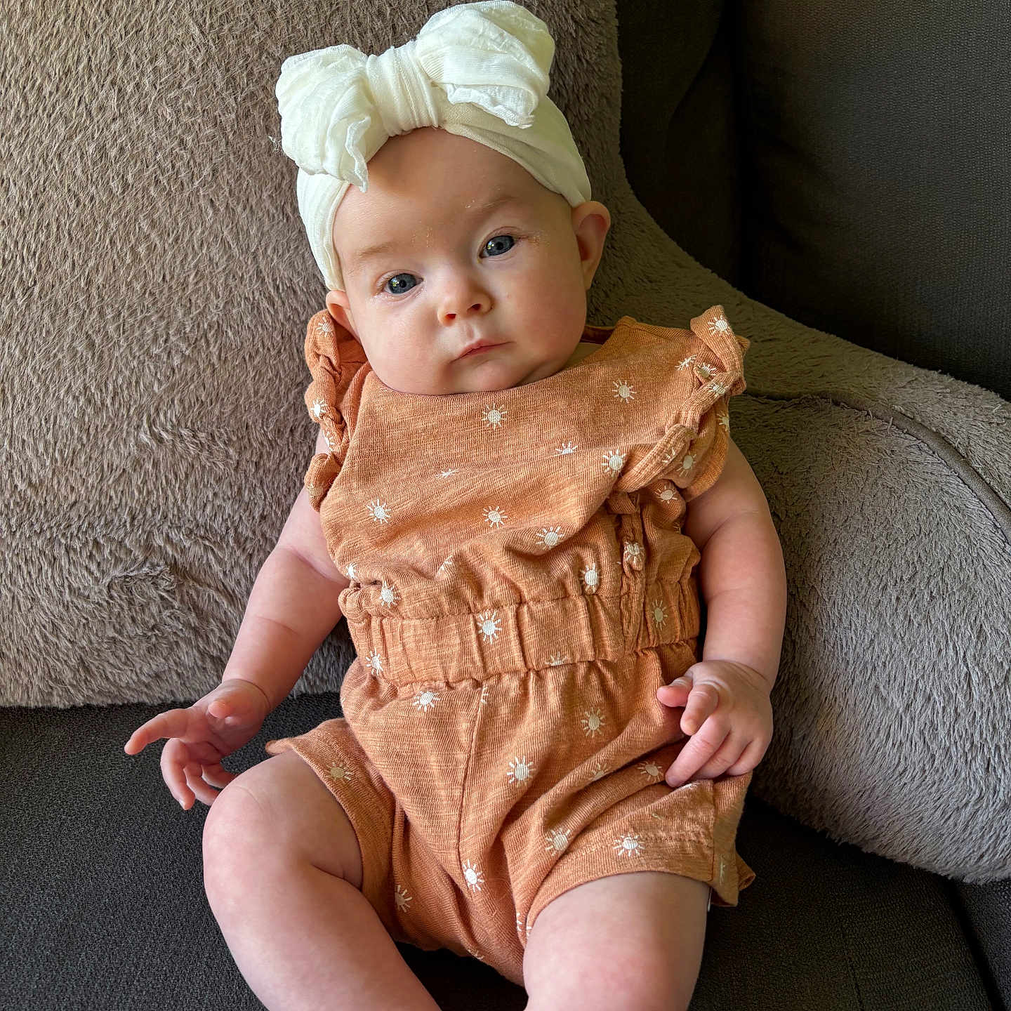 Savannah joined the competition — help win amazing prizes! baby, infant, child, headband, bow, romper, orange_clothing, couch, cushion, soft_texture, seated, indoors, portrait, cute, small_hands, legs, face, expression, looking_at_camera, comfortable