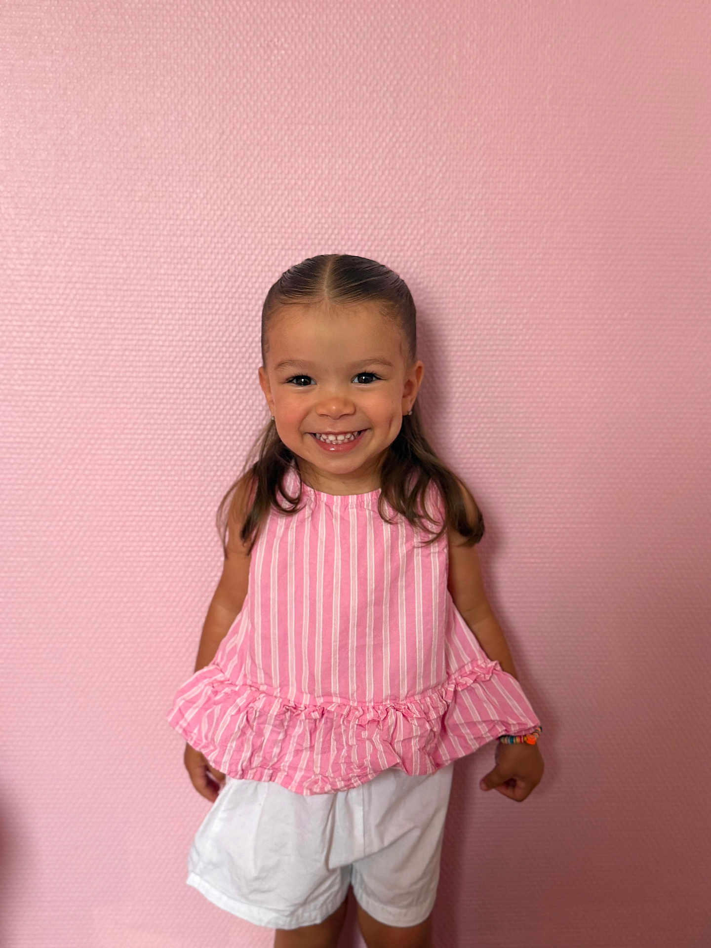 Lila participe au concours pour gagner de l'argent avec cette photo : child, girl, smiling, happy, pink_wall, pink_top, white_shorts, bracelet, dimples, portrait, standing, indoor, cute, fashion, hair, young, childhood, person, casual, colorful