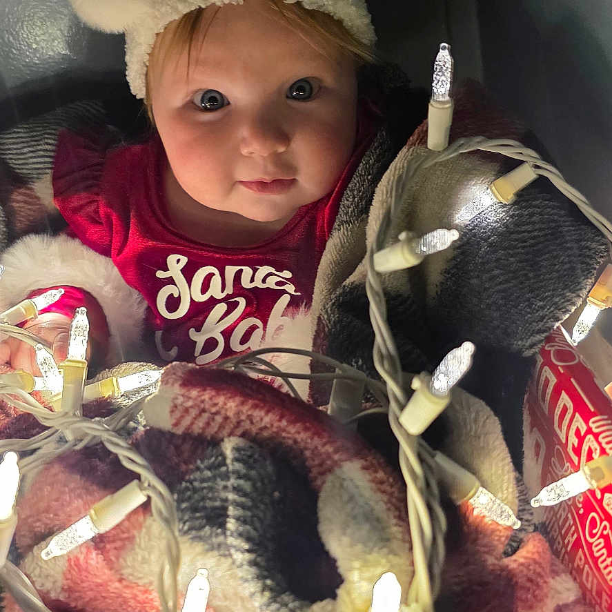 Eleanor is registered to the contest to win money with this photo: baby, blanket, box, celebration, child, christmas, christmas_lights, cozy, cute, decorations, face, festive, holiday, indoors, person, portrait, red_clothing, santa_hat, smile, warm
