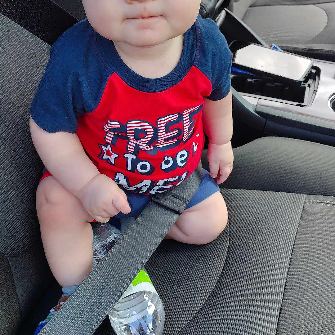Tristan is registered to the contest to win money with this photo: baby, blue_eyes, car_interior, car_seat, child, clothing, cute, expression, indoors, infant, person, portrait, red_shirt, safety, seat, seatbelt, shorts, sitting, vehicle, water_bottle