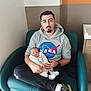 émyjio participe au concours pour gagner de l'argent avec cette photo : man, baby, newborn, hospital, chair, sitting, hoodie, nasa_logo, socks, sleeping, holding, person, indoor, casual_clothing, footwear, furniture, cabinet, watch, facial_hair, relaxing