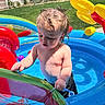 toddler, child, pool, inflatable, water, toy, outdoor, summer, sunlight, grass, playing, wet_hair, backyard, nature, fence, tree, mountain, blue_sky, colorful, fun