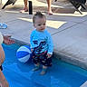 toddler, child, pool, water, beach_ball, swimwear, blue_shirt, dinosaur_print, shorts, adult_hand, lounge_chair, towel, water_bottle, sunlight, outdoor, concrete, summer, hair_spike, person, footwear