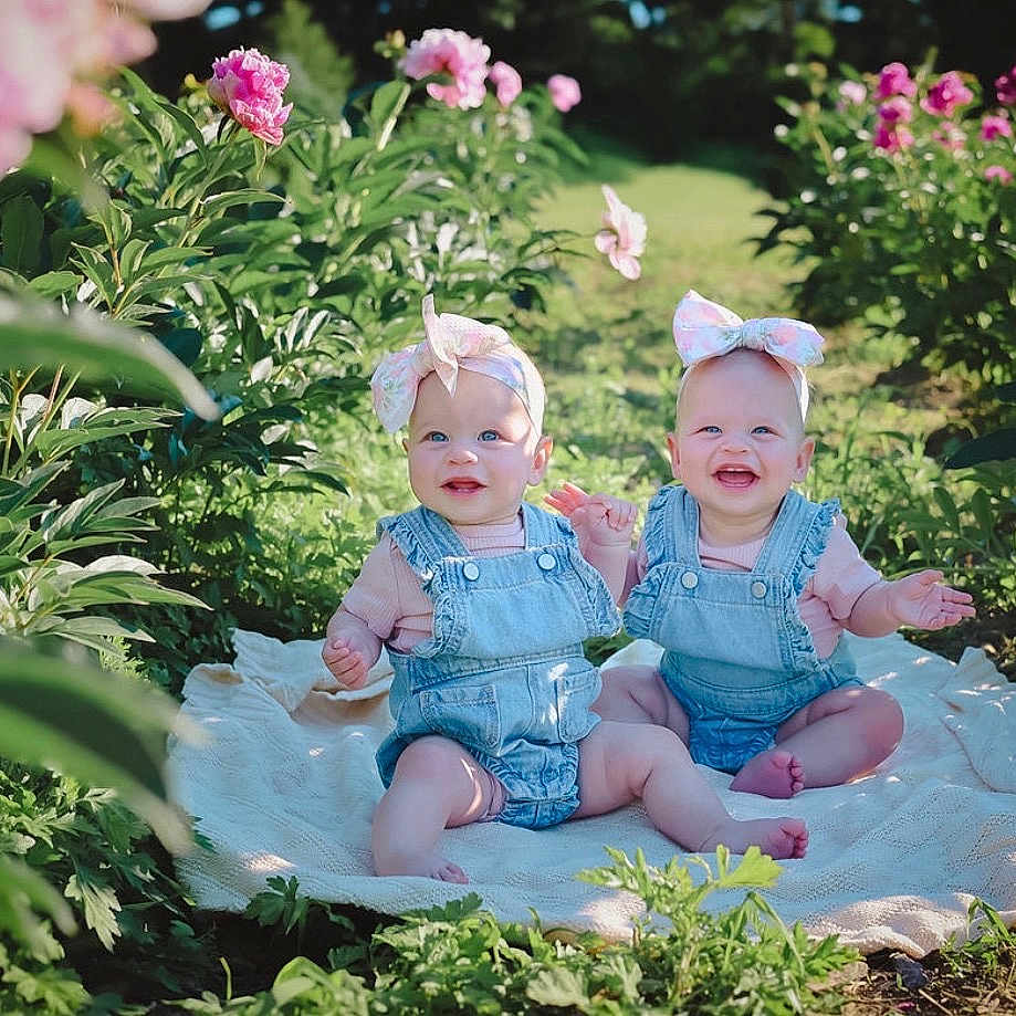 Quinn And Kenna is registered to the contest to win money with this photo: baby, babies, denim_overalls, headband, flower, garden, blanket, outdoor, greenery, plants, smiling, happy, cute, child, nature, sunlight, infant, pink_flowers, sitting, portrait