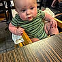 Jaxxon is registered to the contest to win money with this photo: baby, child, high_chair, striped_clothing, green_clothing, restaurant, table, wooden_furniture, curious_expression, drool, indoor, floor_tiles, person, seated, blurry_background, dining_area, chubby_cheeks, hand_on_chair, casual, infant