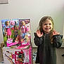 child, girl, smile, toy, barbie, doll, packaging, box, curly_hair, standing, indoor, happy, clothing, floor, tile, play, present, cute, young, person