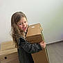 child, smiling, cardboard_box, long_hair, indoor, wooden_chest, floor_tile, happy, girl, clothing, holding, light_wall, curly_hair, person, playful, casual, joy, standing, home, expression