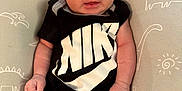 Archer is registered to the contest to win money with this photo: baby, infant, clothing, black_outfit, nike_logo, dinosaur_pattern, pillow, cute, smiling, portrait, indoor, child, newborn, person, human, skin, face, hand, sitting, young