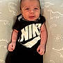 baby, infant, clothing, black_outfit, nike_logo, dinosaur_pattern, pillow, cute, smiling, portrait, indoor, child, newborn, person, human, skin, face, hand, sitting, young
