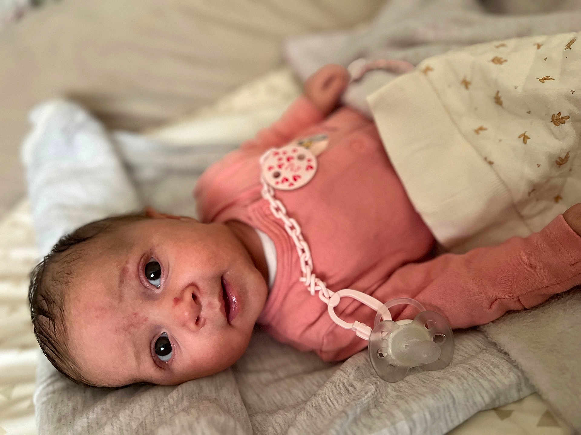 Assil a rejoint le concours — aidez-le/la à gagner de superbes lots ! baby, infant, newborn, pacifier, pacifier_clip, pacifier_chain, onesie, pink_clothing, blanket, bedding, lying_down, portrait, close_up, big_eyes, gaze, adorable, cozy, soft_texture, sleeping_area, skin