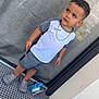 bottle, boy, casual, chain_necklace, child, concrete_floor, crocs, curly_hair, expression, glass_door, gray_clothing, outdoor, reflection, short_sleeves, shorts, side_view, standing, toddler, toy_helicopter, trash_bag