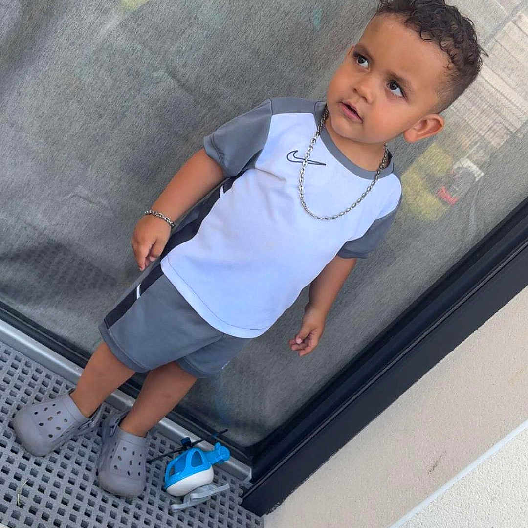 Swaïn a rejoint le concours — aidez-le/la à gagner de superbes lots ! bottle, boy, casual, chain_necklace, child, concrete_floor, crocs, curly_hair, expression, glass_door, gray_clothing, outdoor, reflection, short_sleeves, shorts, side_view, standing, toddler, toy_helicopter, trash_bag