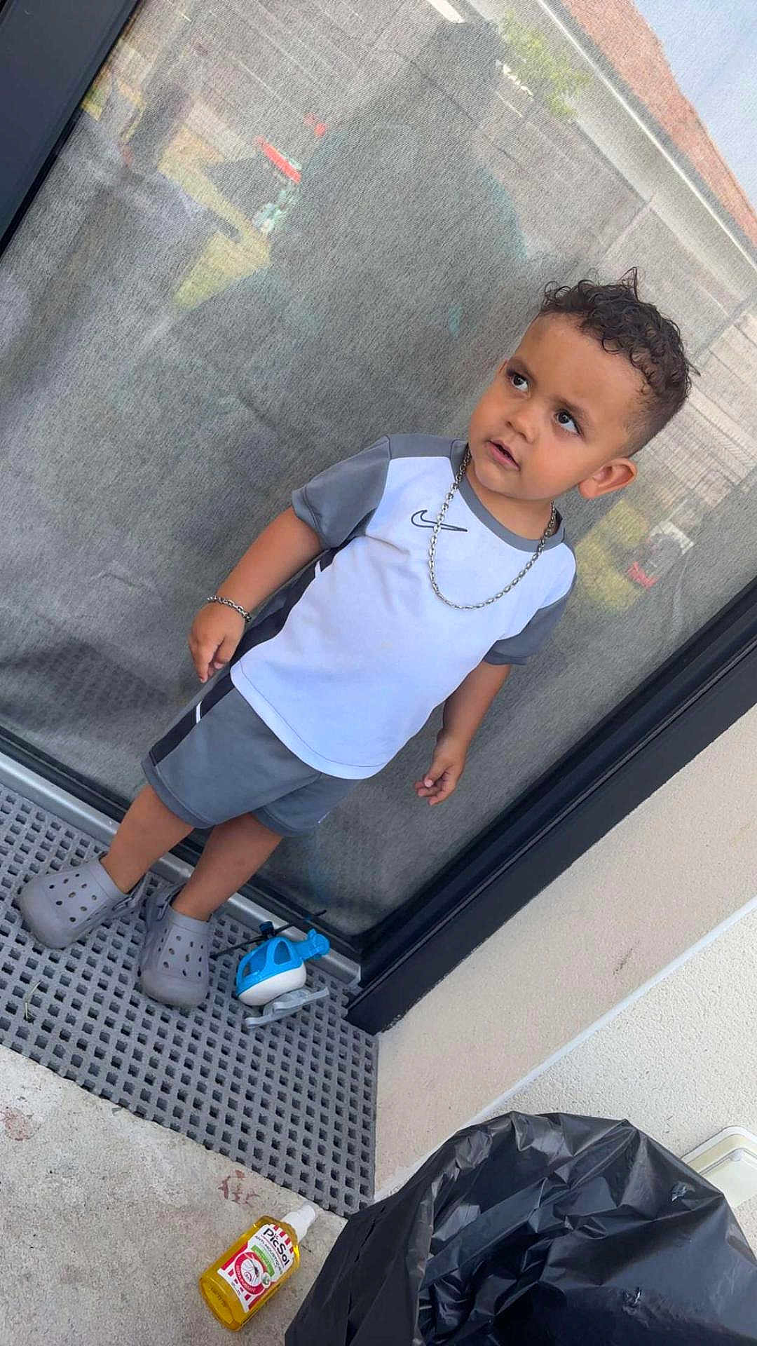 Swaïn a rejoint le concours — aidez-le/la à gagner de superbes lots ! toddler, child, boy, curly_hair, gray_clothing, crocs, chain_necklace, toy_helicopter, glass_door, outdoor, concrete_floor, trash_bag, bottle, side_view, casual, shorts, short_sleeves, standing, expression, reflection