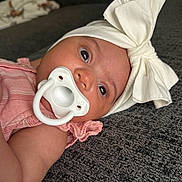 Rayah joined the competition — help win amazing prizes! baby, infant, pacifier, headband, bow, pink_clothing, couch, gray, texture, closeup, face, eyes, skin, child, indoor, portrait, cute, sleepwear, relaxing, lying_down