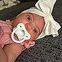 baby, infant, pacifier, headband, bow, pink_clothing, couch, gray, texture, closeup, face, eyes, skin, child, indoor, portrait, cute, sleepwear, relaxing, lying_down