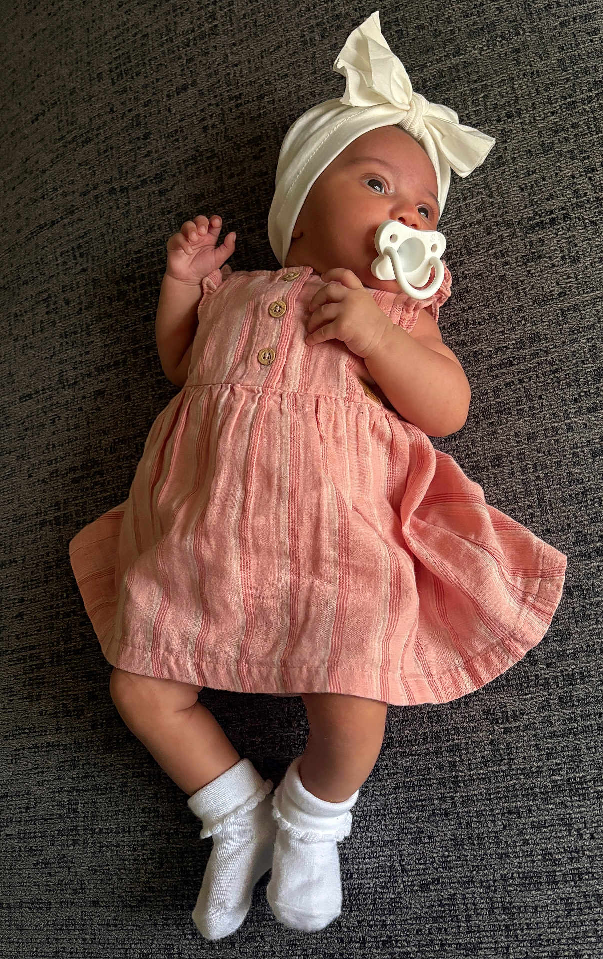 Rayah is registered to the contest to win money with this photo: baby, infant, pacifier, pink_dress, headband, bow, white_socks, carpet, textured_background, wooden_buttons, clothing, lying_down, child, cute, adorable, small_hands, small_feet, portrait, indoor, calm