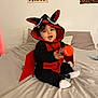 baby, child, costume, bat, red, black, bed, pillow, smiling, indoors, toy, cute, wall_art, happy, sitting, clothing, footwear, headwear, person, infant