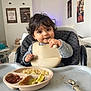 Kamil a rejoint le concours — aidez-le/la à gagner de superbes lots ! toddler, child, high_chair, bib, spoon, food, meal, plate, indoor, curly_hair, cute, happy, table, dining, home, wall_art, poster, furniture, person, eating