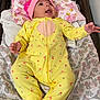 baby, infant, yellow_onesie, pink_hat, lying_down, blanket, patterned_blanket, cushion, indoor, child, cute, happy, smiling, person, small_hand, footed_pajamas, headwear, floral_pattern, soft_fabric, cozy