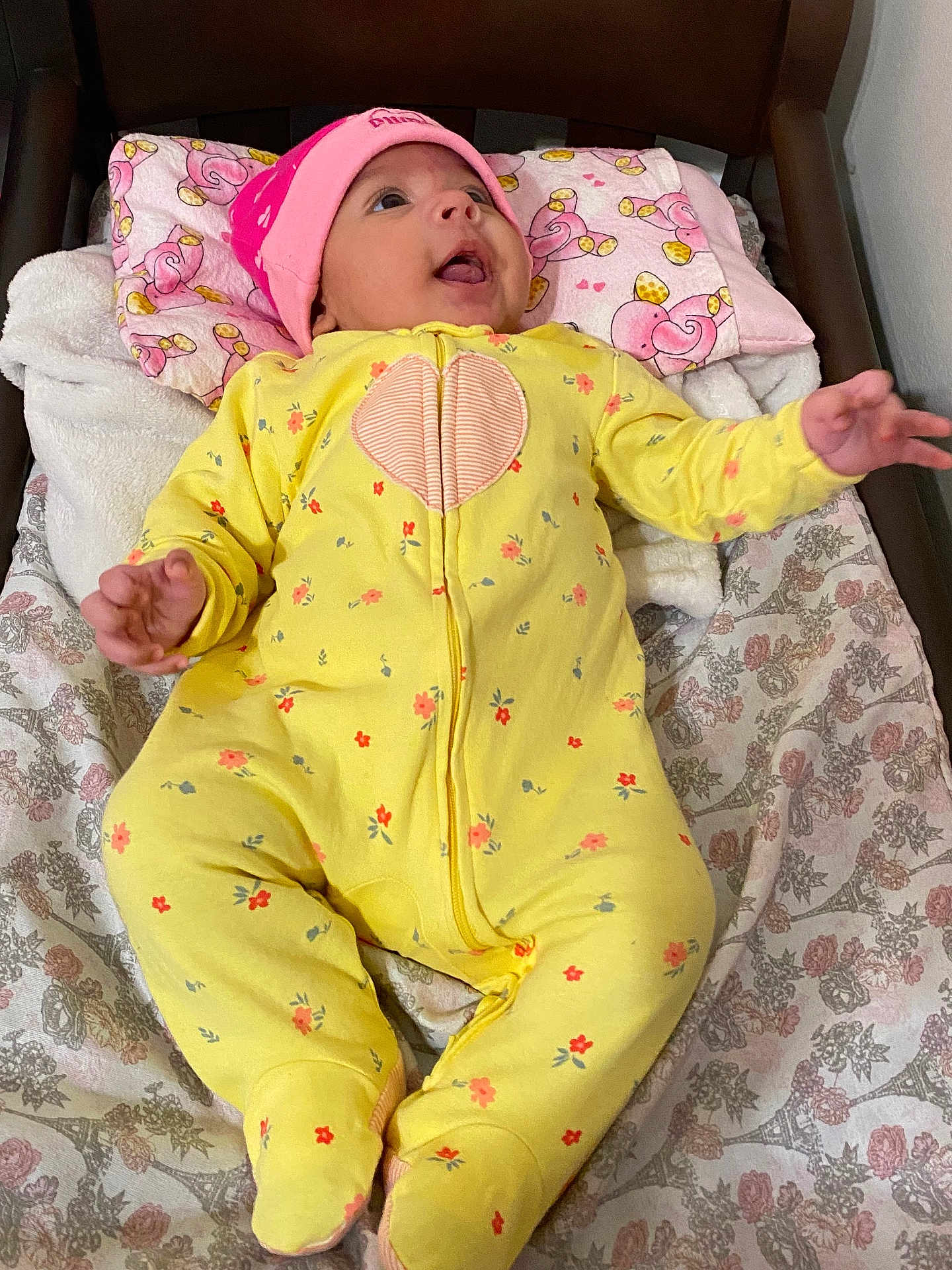 Emmy joined the competition — help win amazing prizes! baby, infant, yellow_onesie, pink_hat, lying_down, blanket, patterned_blanket, cushion, indoor, child, cute, happy, smiling, person, small_hand, footed_pajamas, headwear, floral_pattern, soft_fabric, cozy