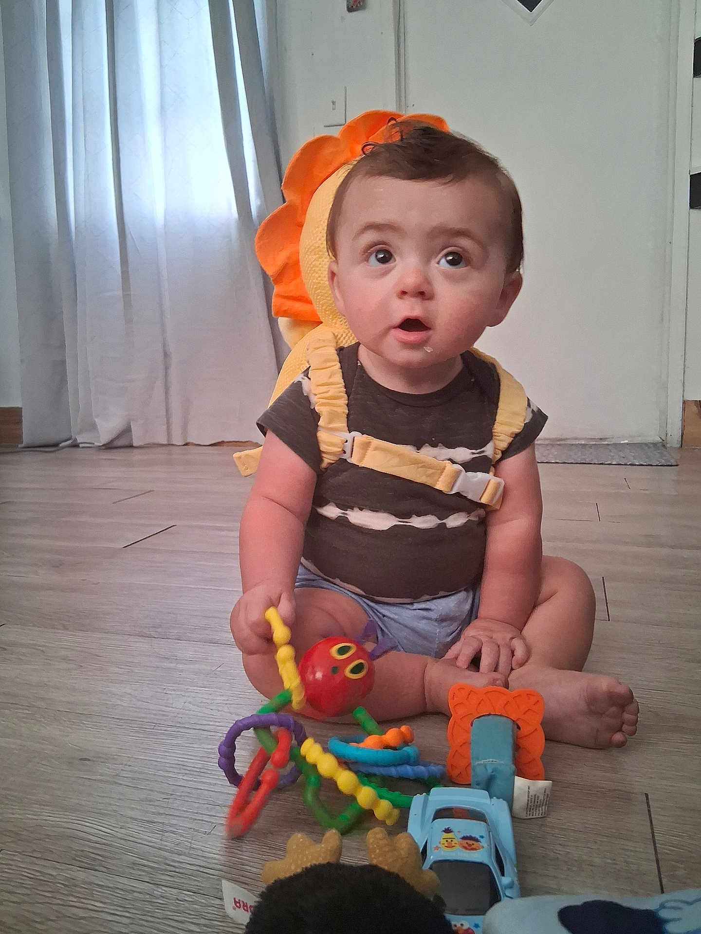 Colton is registered to the contest to win money with this photo: baby, backpack, blue, child, clothing, curtains, cute, expression, floor, hand, indoor, octopus_toy, orange, person, playful, sitting, toy, wide_eyes, wooden_floor, yellow