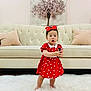 Esol is registered to the contest to win money with this photo: child, toddler, red_dress, polka_dot, headband, barefoot, white_rug, sofa, cushion, painting, tree, indoor, person, cute, standing, expression, furniture, carpet, holding_object, home