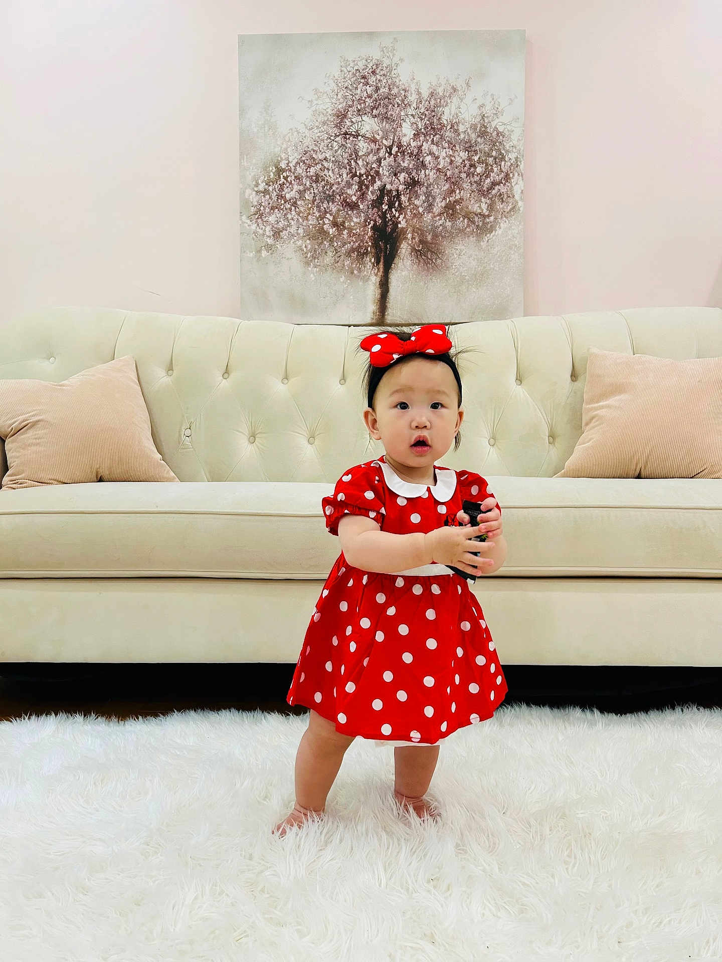 Esol is registered to the contest to win money with this photo: child, toddler, red_dress, polka_dot, headband, barefoot, white_rug, sofa, cushion, painting, tree, indoor, person, cute, standing, expression, furniture, carpet, holding_object, home