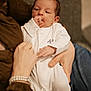 Aloïs participe au concours pour gagner de l'argent avec cette photo : baby, infant, white_onesie, hand, thumb_sucking, adult_hand, holding, couch, fabric, jeans, brown_shirt, bracelet, indoor, portrait, cute, calm, closeup, person, newborn, comfort