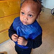 Khari joined the competition — help win amazing prizes! toddler, child, indoor, blue_bib, black_clothing, curly_hair, floor, cabinet, shoes, person, looking_up, curious, young_child, small_hands, standing, home, expression, hair_twists, casual, floor_tiles