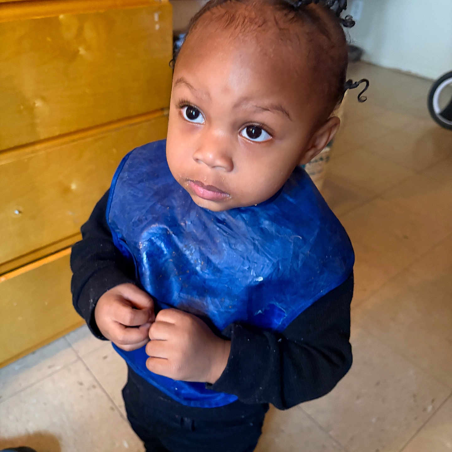 Khari joined the competition — help win amazing prizes! toddler, child, indoor, blue_bib, black_clothing, curly_hair, floor, cabinet, shoes, person, looking_up, curious, young_child, small_hands, standing, home, expression, hair_twists, casual, floor_tiles