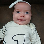 Willow is registered to the contest to win money with this photo: baby, headband, bow, heart_pattern, clothing, smile, tongue, face, infant, cute, child, portrait, sitting, white, cream, black, background, closeup, happy, person
