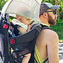 baby, backpack, beard, carrier, casual_clothing, child, footwear, greenery, headwear, man, nature, outdoor, park, person, shade, summer, sunglasses, sunlight, trees, walking