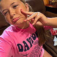 Mikayla is registered to the contest to win money with this photo: girl, child, pink_shirt, turtle_design, pouty_face, hand_gesture, long_hair, couch, living_room, lamp, earrings, casual_clothing, indoor, seated, brown_furniture, window, curtains, relaxed, playful, teenager