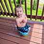 child, boy, bucket, wet_hair, smiling, outdoor, deck, wooden_floor, sunlight, greenery, playful, summer, bare_chest, shorts, grass, railing, happy, sitting, water, daylight