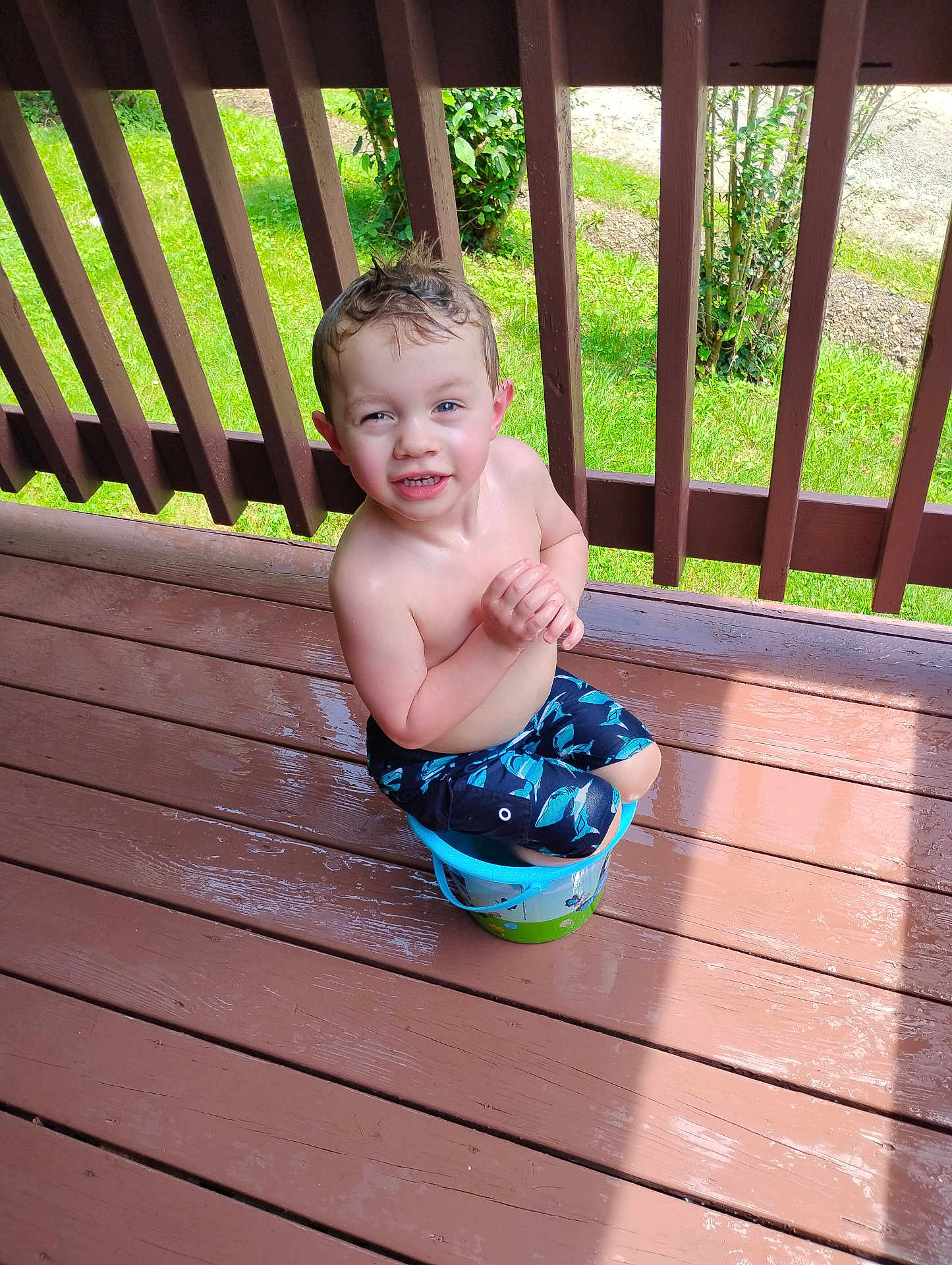 Gunner joined the competition — help win amazing prizes! child, boy, bucket, wet_hair, smiling, outdoor, deck, wooden_floor, sunlight, greenery, playful, summer, bare_chest, shorts, grass, railing, happy, sitting, water, daylight