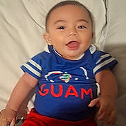 Denario is registered to the contest to win money with this photo: baby, child, infant, smiling, happy, portrait, indoors, bed, pillow, blue_shirt, red_pants, clothing, sitting, lying_down, face, eyes, mouth, hands, bedroom, guam