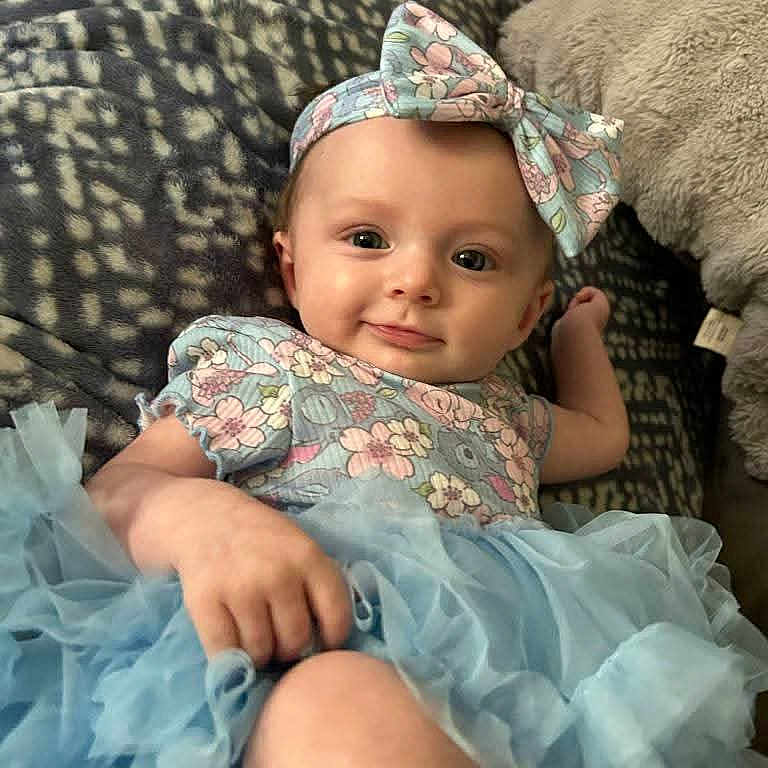 Paige joined the competition — help win amazing prizes! baby, blanket, blue_tutu, bow, child, cuddly, cute, face, floral_top, hand, happy, headband, indoors, infant, lying_down, patterned, portrait, skin, smiling, soft_toys