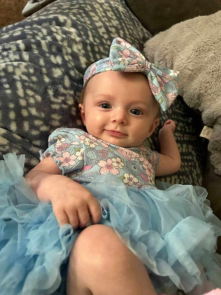 Paige joined the competition — help win amazing prizes! baby, child, smiling, headband, bow, blue_tutu, floral_top, cute, lying_down, blanket, patterned, infant, happy, portrait, indoors, soft_toys, cuddly, hand, face, skin