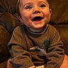 baby, brown_couch, casual_clothing, child, cozy, cute, cute_expression, face, feet, hands, happy, indoor, infant, person, portrait, seated, smiling, sweater, warm_lighting, young_child