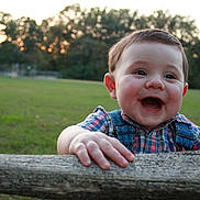 Winston is registered to the contest to win money with this photo: baby, child, smiling, happy, outdoor, park, grass, trees, sunset, portrait, face, eyes, mouth, hands, wood, log, plaid_shirt, overalls, baby_boy, nature