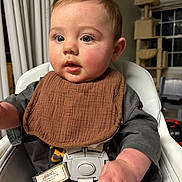 Winston joined the competition — help win amazing prizes! baby, child, high_chair, infant, bib, sitting, indoors, portrait, face, eyes, brown_bib, gray_clothing, happy, curious, open_mouth, short_hair, home, curtains, cat_tree, window