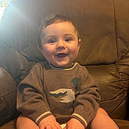 Winston is registered to the contest to win money with this photo: baby, child, smiling, sweater, duck_design, brown_leather_chair, happy, hands, feet, indoors, portrait, cute, sitting, young_child, cozy, warm_lighting, skin, face, eyes, hair