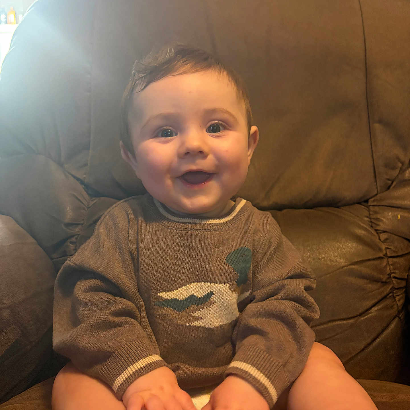 Winston is registered to the contest to win money with this photo: baby, child, smiling, sweater, duck_design, brown_leather_chair, happy, hands, feet, indoors, portrait, cute, sitting, young_child, cozy, warm_lighting, skin, face, eyes, hair